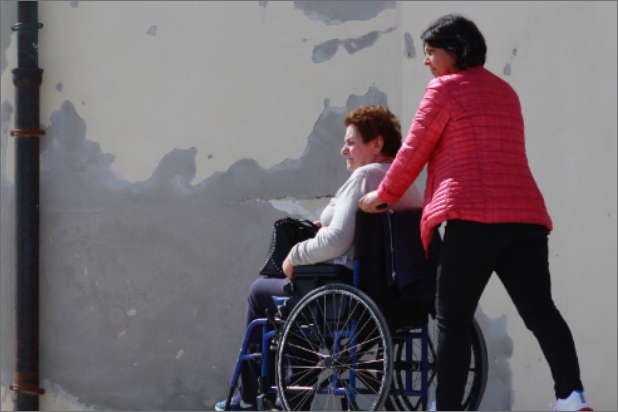 Kobieta prowadzi inną kobietę na wózku, w tle szara ściana budynku.