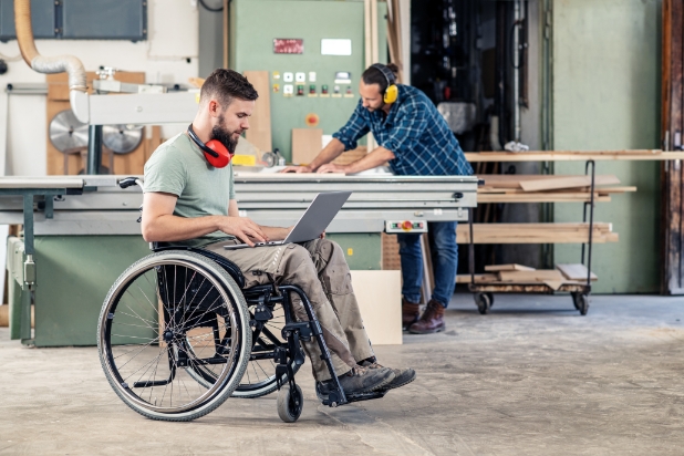 Na pierwszym planie mężczyzna na wózku, na kolanach ma laptop. Na drugim planie mężczyzna pracujący przy maszynie przemysłowej.