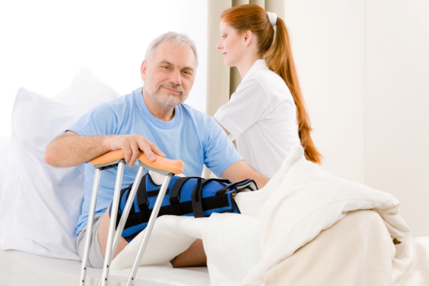 Starszy mężczyzna siedzi na łóżku, ma ortezę na nodze, na drugim planie obok niego stoi pielęgniarka.