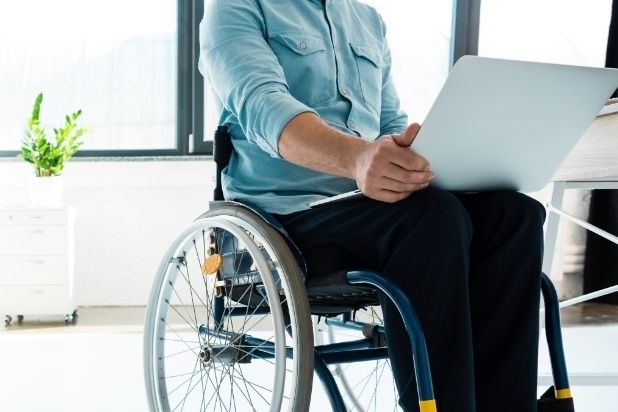 Fragmentaryczna postać mężczyzny na wózku, mężczyzna w stroju biurowym, na kolanach trzyma laptop.