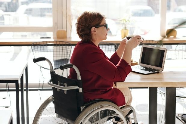 Kobieta na wózku, z kubkiem w dłoniach. Pracuje pracuje przy biurku na laptopie.