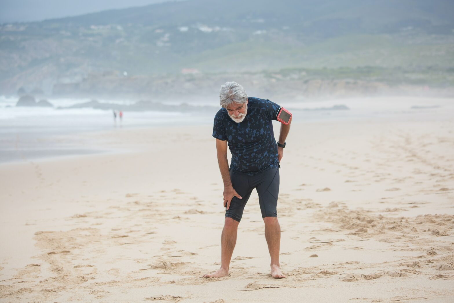 Mężczyzna na plaży pochylający się do przodu z miną sugerującą ból, trzyma się za plecy.