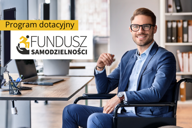 Młody uśmiechnięty mężczyzna w okularach i garniturze siedzi na wózku przy biurku, na którym jest laptop. W lewym górnym rogu czarny napis na żółtej belce: Program dotacyjny, a pod spodem logo Funduszu Samodzielności