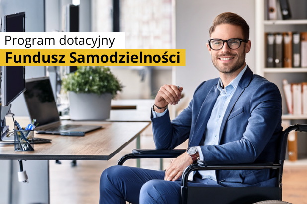 Młody uśmiechnięty mężczyzna w okularach i garniturze siedzi na wózku przy biurku, na którym jest laptop. W lewym górnym rogu czarny napis na białej i żółtej belce: Program dotacyjny Fundusz Samodzielności.