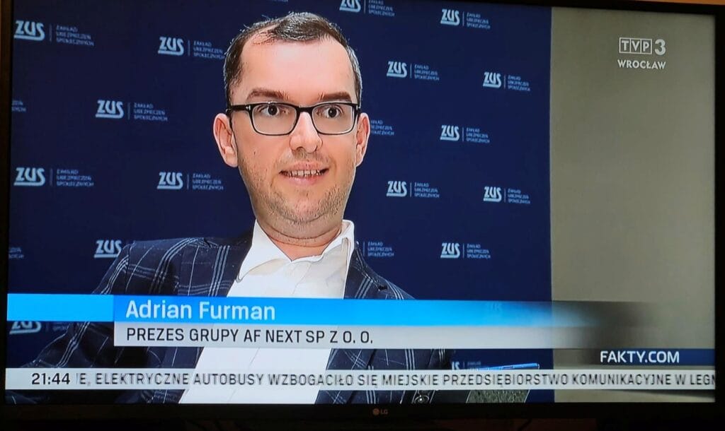 Adrian Furman w okularach, ciemnej marynarce i białej koszuli na ekranie TVP3 Wrocław, w tle plansza z logo Zakładu Ubezpieczeń Społecznych.
