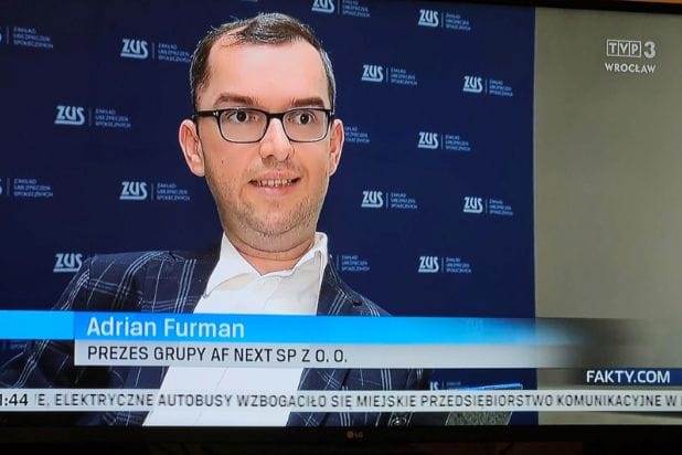 Adrian Furman w okularach, ciemnej marynarce i białej koszuli na ekranie TVP3 Wrocław, w tle plansza z logo Zakładu Ubezpieczeń Społecznych.