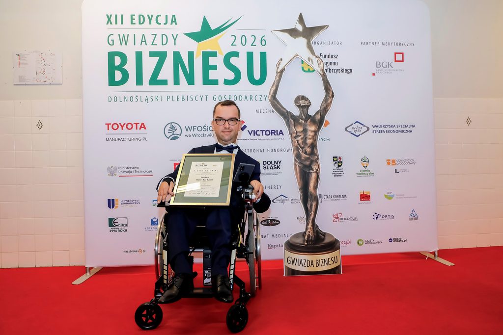Adrian Furman w uroczystym galowym garniturze na wózku z Certyfikatem nagrody dla Fundacji Biznes Bez Barier. Obok statuetka. W tle ścianka z logotypami partnerów Plebiscytu.