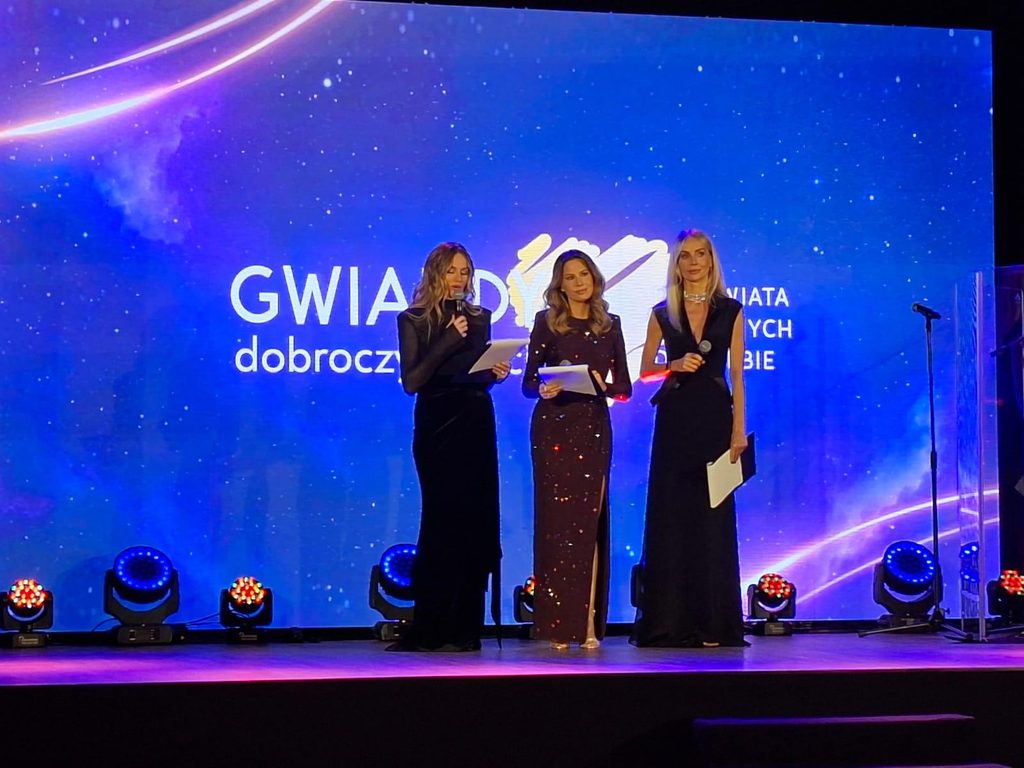 Trzy kobiety w strojach galowych/wieczorowych na scenie podczas części oficjalnej