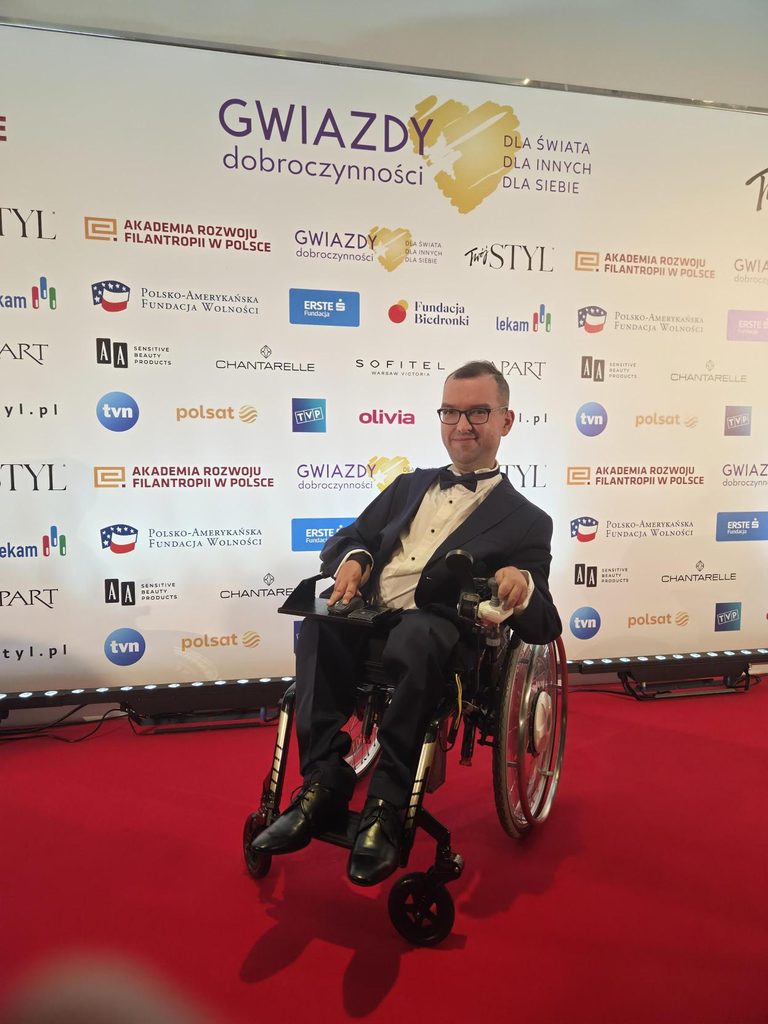 Adrian Furman na wózku w ciemnym garniturze i z ciemną muszką przed ścianką z logotypami partnerów wydarzenia