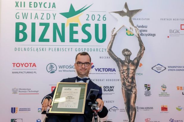 Adrian Furman w uroczystym galowym garniturze na wózku z Certyfikatem nagrody dla Fundacji Biznes Bez Barier. Obok statuetka. W tle ścianka z logotypami partnerów Plebiscytu.